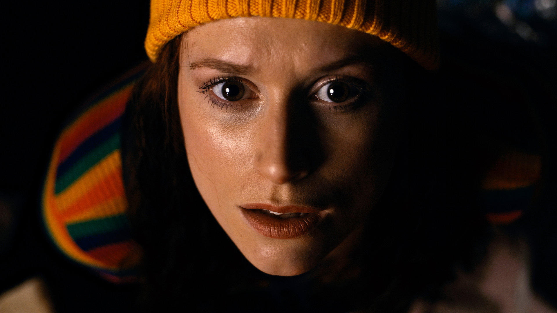DISSOCIATION. Actor Angela Leta Kaye as the Genealogist. She is a young woman with auburn hair, a yellow beanie, and a rainbow long-sleeve shirt. In this image, she stares directly at the camera, in horror of what she's seeing off-screen.