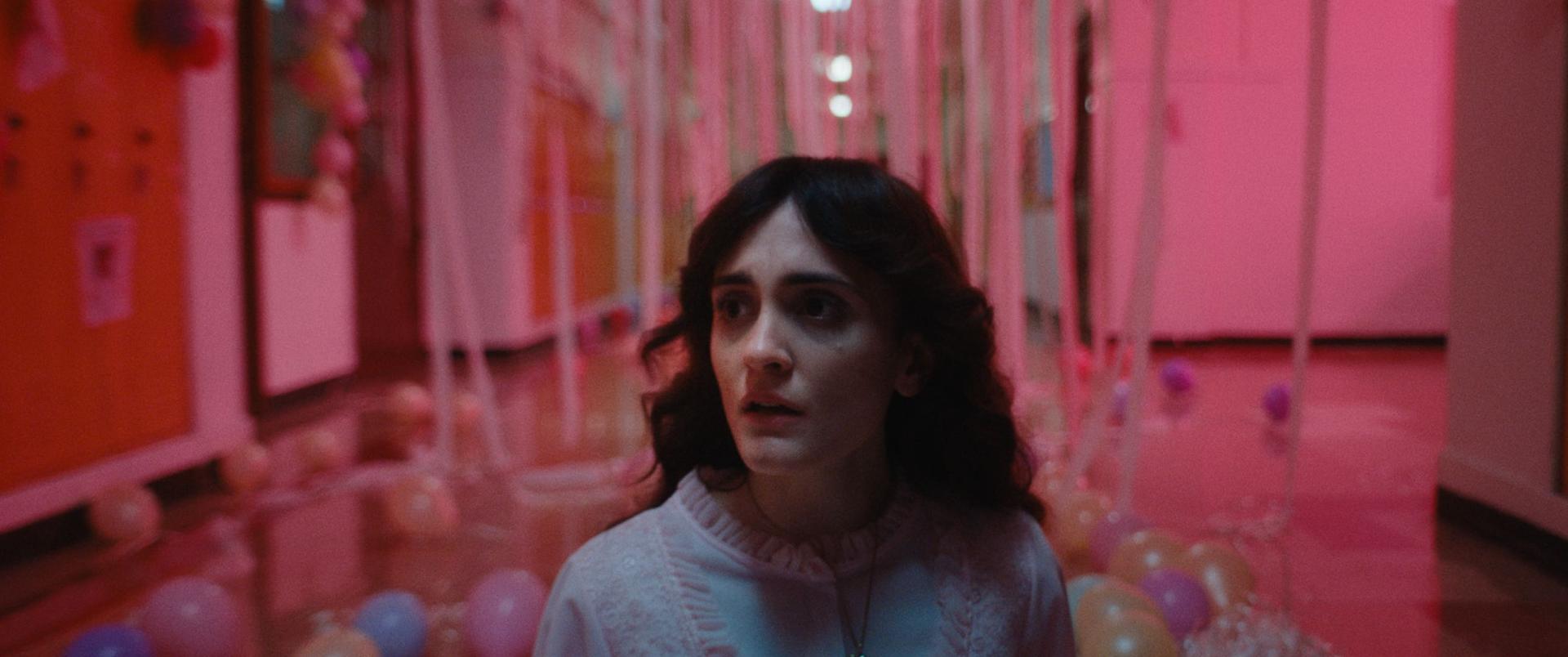 A brunette teen girl in a nightgown looks frightened in an eerie hallway surrounded by pink lighting and prom old balloons and streamers.