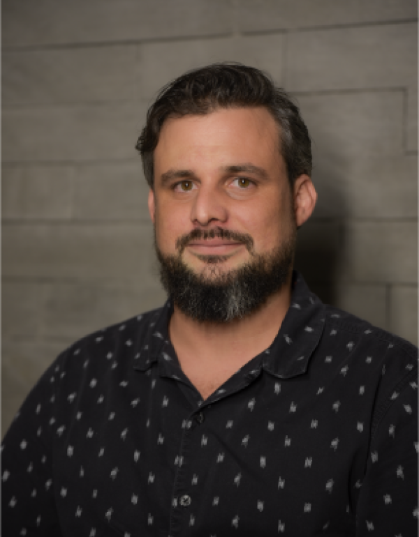 A man with medium-length dark hair and a beard is facing the camera and smiling softly. He is wearing a dark blue shirt. The lighting is warm and even, showing his face clearly.