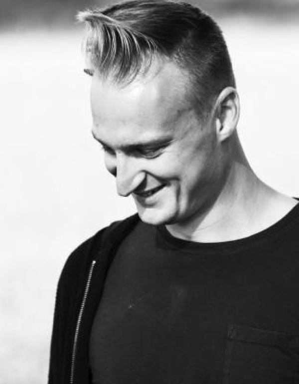 Black and white mid-shot of a smiling Caucasian man with blonde hair cut short at the sides and styled in a quiff at the top wearing a black t-shirt. He stands in a 45 degree left side profile with his head slightly angled down towards the floor.
