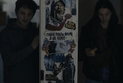 A dimly lit scene shows a young man and a young woman standing on either side of a door covered in collage-style posters. The posters feature distressed faces and bold text in Spanish, including “¿Quién puede matar a un niño?” The man, wearing a hoodie, looks tense as he touches the door, while the woman looks down at something in her hands. The atmosphere feels dark and suspenseful.