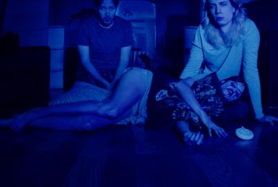 Two seated figures stare straight ahead in a dark, blue room. Another is lying down beside them... bloody and unwell.