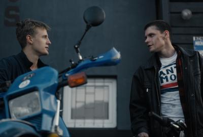Two young men talking next to a blue motorcycle and black bicycle.