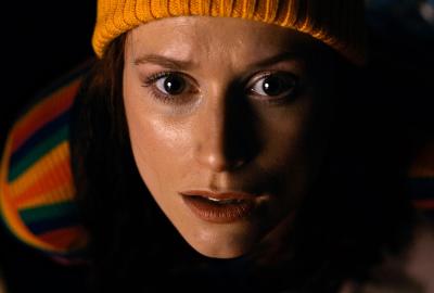 DISSOCIATION. Actor Angela Leta Kaye as the Genealogist. She is a young woman with auburn hair, a yellow beanie, and a rainbow long-sleeve shirt. In this image, she stares directly at the camera, in horror of what she's seeing off-screen.