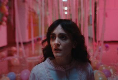 A brunette teen girl in a nightgown looks frightened in an eerie hallway surrounded by pink lighting and prom old balloons and streamers.