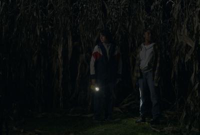 Cole and Shelby stand still in the middle of a cornfield, their flashlights illuminating the stalks around them.