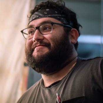 A smiling bearded man wearing glasses and a black bandana looks off-camera in a candid close-up.